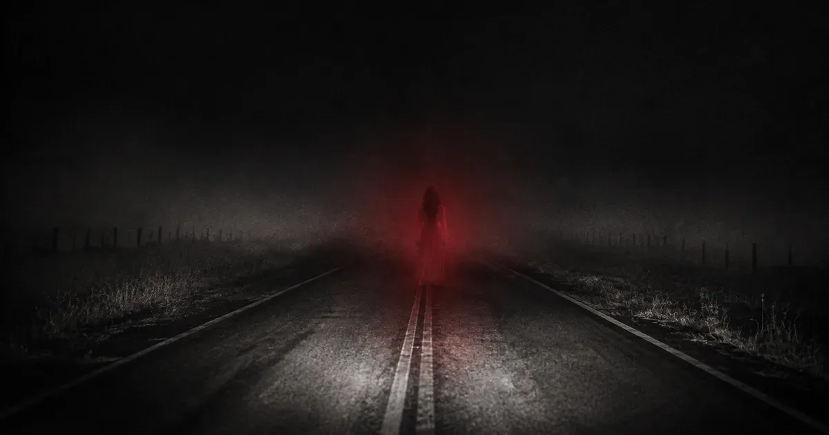 Una ruta solitaria de noche entre la niebla, con la silueta difusa de una mujer de blanco parada sobre el asfalto a la distancia, iluminada por un tenue resplandor rojo