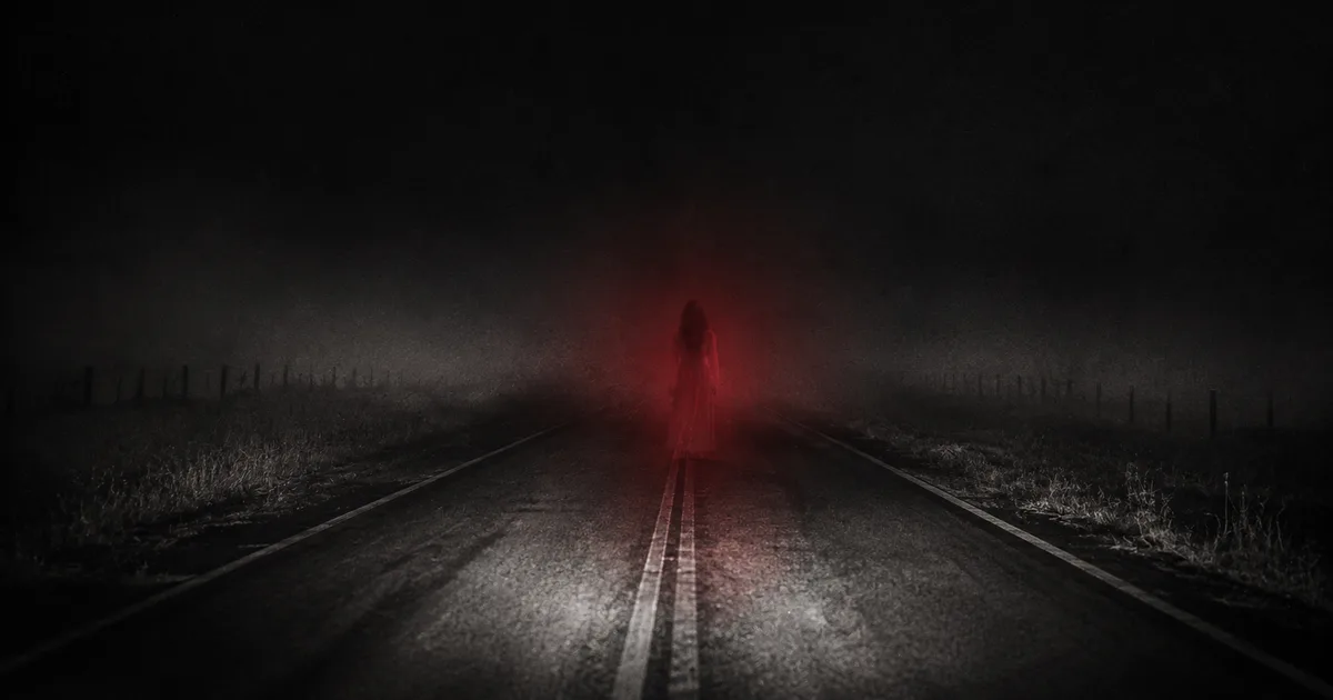 Una ruta solitaria de noche entre la niebla, con la silueta difusa de una mujer de blanco parada sobre el asfalto a la distancia, iluminada por un tenue resplandor rojo